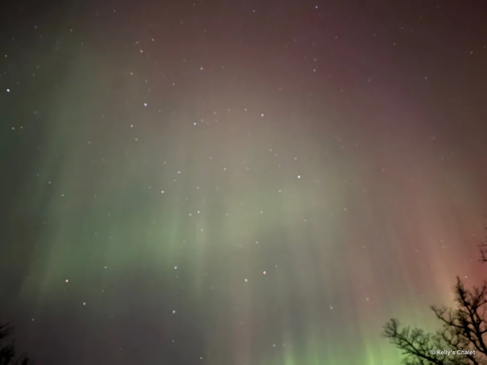 Pink and green aurora curtains above treetops near Grand Beach, photographed away from Kelly’s Chalet