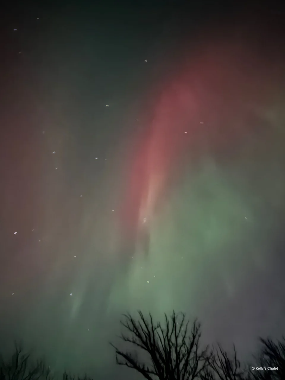 A red aurora ribbon and tree silhouettes in the Grand Beach area, photographed away from Kelly’s Chalet