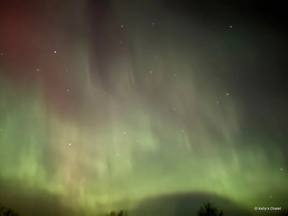 Soft green and pink aurora glow above treetops near Grand Beach, photographed away from Kelly’s Chalet